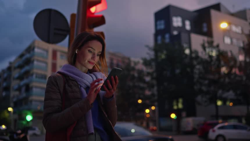 Woman using a smartphone at an urban intersection at dusk with a red traffic light in the background