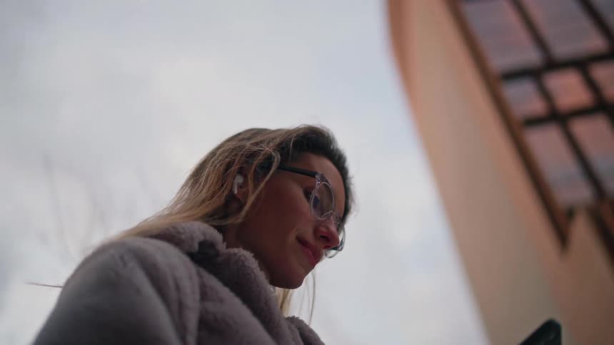 A woman with long brown hair wearing a purple fleece coat and wireless earbuds uses her phone while walking through a city with modern glass buildings during the evening