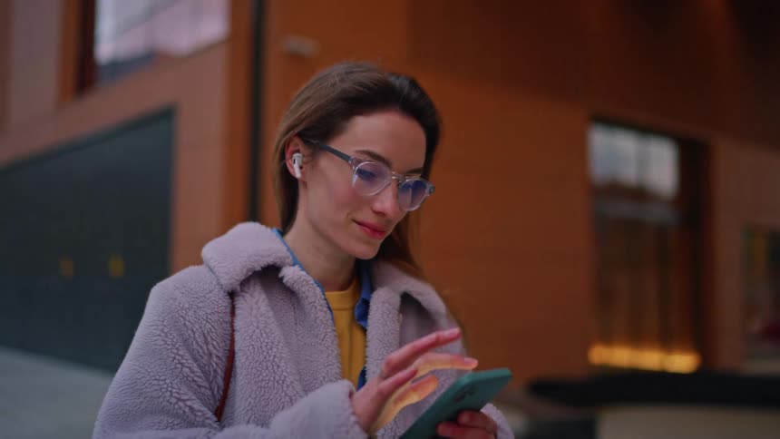 On a modern city street a woman in a plush lightgray coat and clear glasses wearing wireless earbuds smiles while tapping a teal smartphone soft morning light and blurred buildings framing the scene