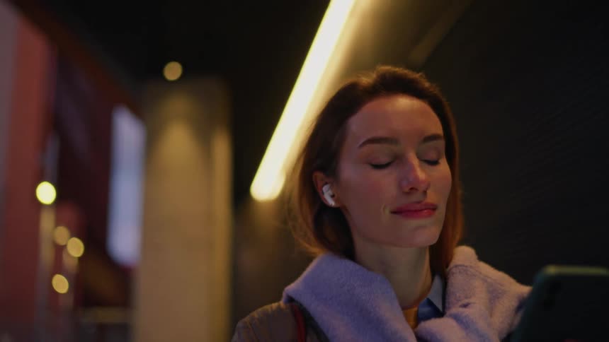 Woman smiles and uses her smartphone while walking close to an urban building at night