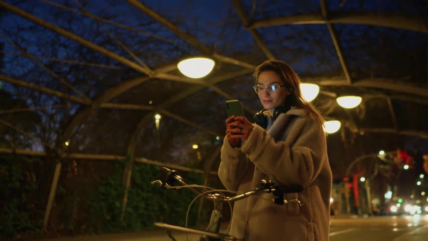 At night under an illuminated arched walkway a woman in a plush light gray coat and glasses stops beside a bicycle smiling at a smartphone as warm lights glow from headlights