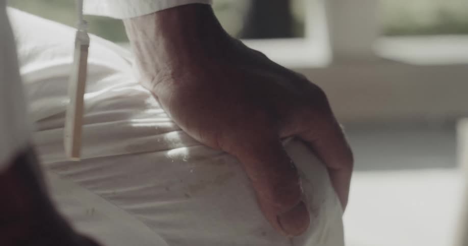 A close view shows the wrinkled hand of a person resting on a white fabric surface while a small wooden tag on a white string swings gently back and forth