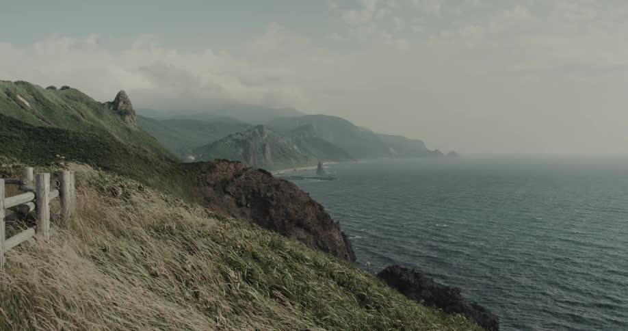 A misty coastal cliffside with a wooden fence overlooks the sea under a soft cloudy sky