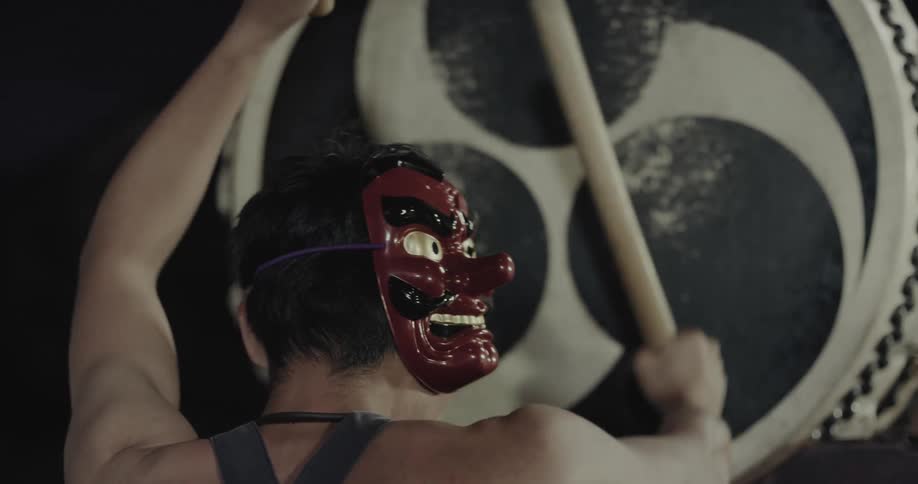 A man wearing a red demonic mask on the back of his head plays a large taiko drum with wooden sticks he strikes the drum forcefully while turning his body showing the mask and his muscular back against a dark background