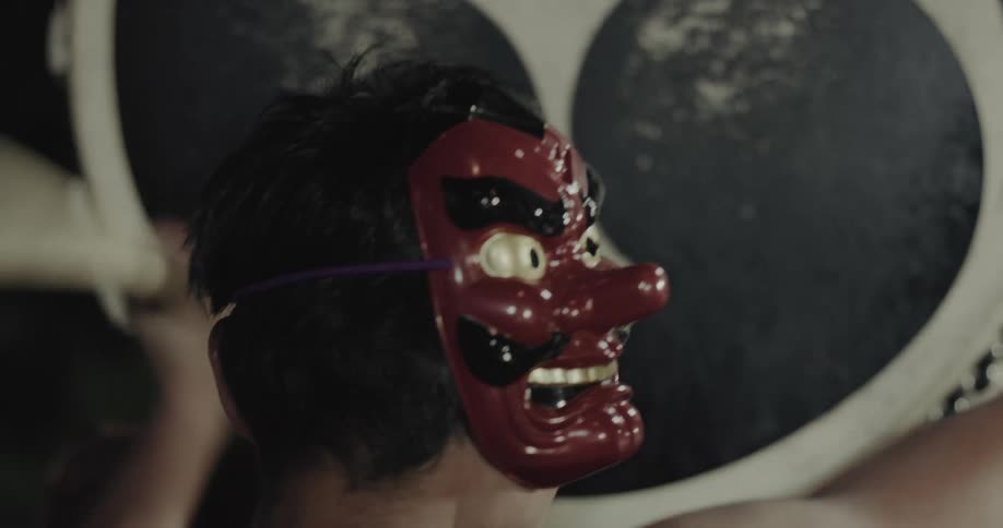 A drummer wearing a red longnosed tengu mask on the side of his head vigorously strikes a large taiko drum decorated with a mitsutomoe symbol using thick wooden bachi sticks during a traditional performance