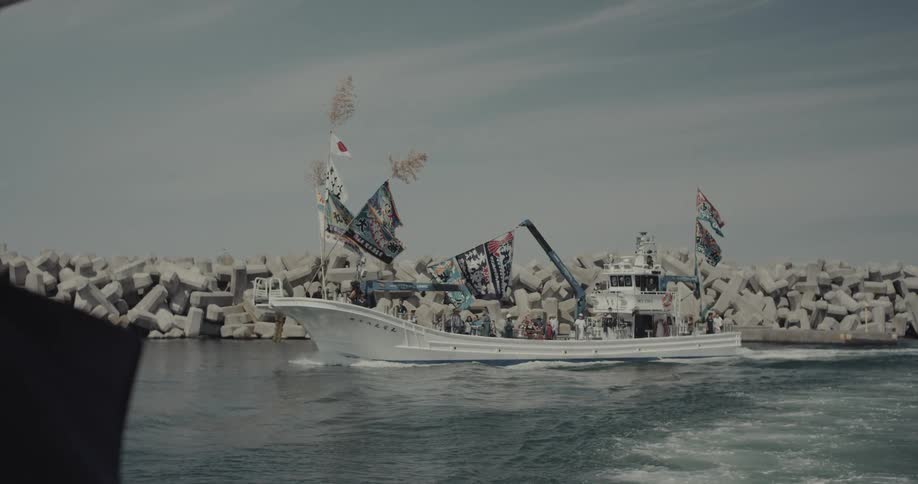 A white fishing boat decorated with numerous traditional colorful banners and japanese flags carries a large group of people across the water past a concrete tetrapod breakwater while a bald man appears in the foreground