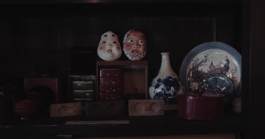 A collection of traditional japanese artifacts and masks rests on a dark wooden shelf the dimly lit scene features an okame mask a hyottoko mask and intricately patterned ceramics
