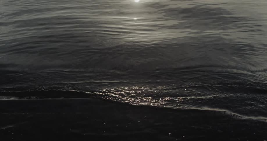 Dark rhythmic ocean ripples flow across the frame while bright sunlight reflects off the moving surface creating a shimmering effect on the deeptoned water