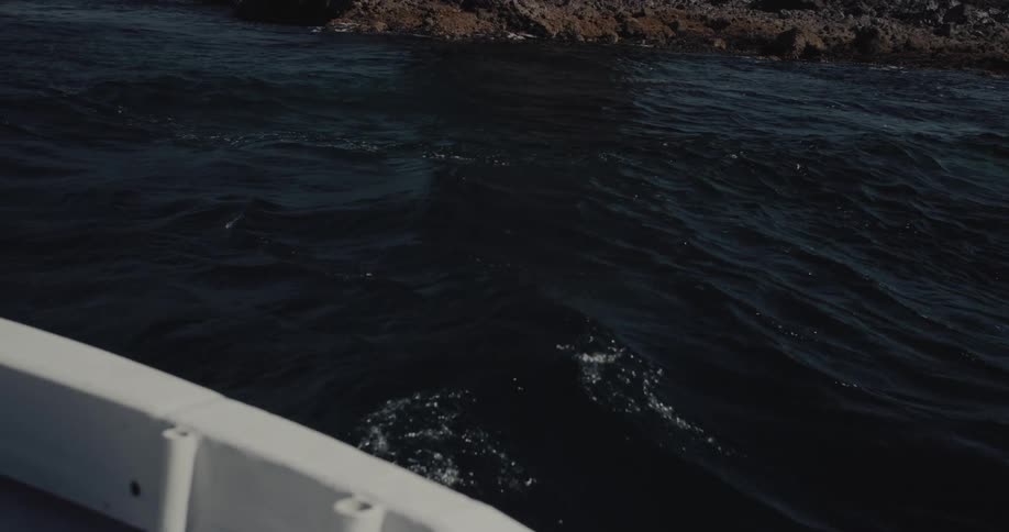 A white boat cruises across deep blue coastal waters providing a close view of a massive dark rock formation rising from the sea alongside a rugged shoreline backed by steep verdant mountain ranges under a clear sky