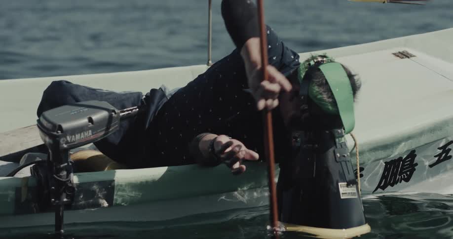 A person in a small boat wearing a mask like apparatus is leaning over the side holding a long wooden pole in the water fishing