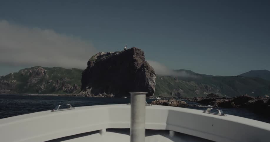 A boat approaches a rugged dark rock formation with a small lighthouse on top set against a backdrop of green hilly coastline under a partly cloudy blue sky