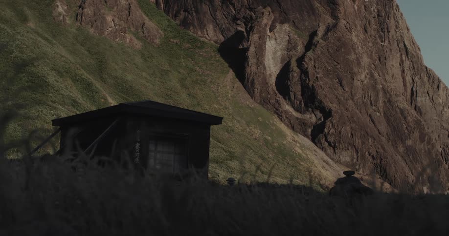 Small building at the foot of a mountain as plants move in the wind