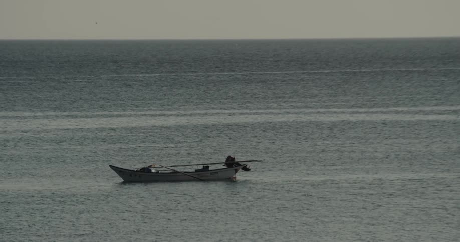 A solitary fishing boat floats adrift on the vast metallic expanse of the open sea bobbing gently with the current under a hazy sky