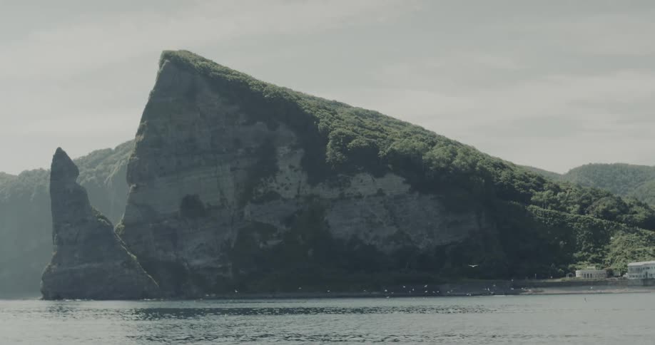 A large mountainous rock rising from the ocean at midday its surface covered in lush green vegetation as sunlight highlights its rugged shape