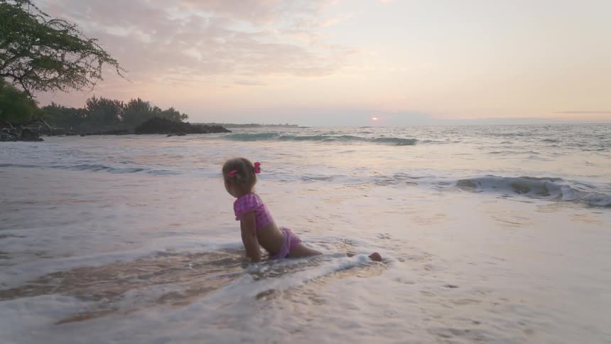 A young girl in a swimsuit lying on the beach at sunset as gentle waves wash over her then standing up and running out of the water with excitement