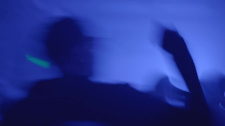 A crowd of people dances in a chaotic blur of motion as strobing blue red and purple lights sweep across the hazy nightclub