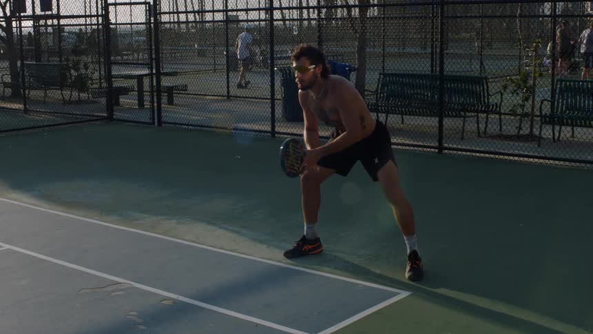 Player holds the racket with both hands in a ready stance on a fenced outdoor sports court