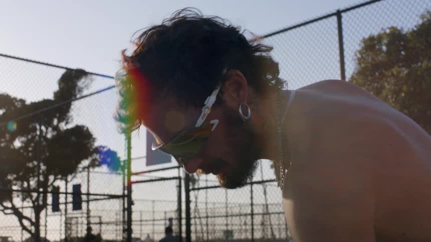 Young man with facial hair wearing sunglasses a necklace and earrings standing shirtless in a sports yard at sunset