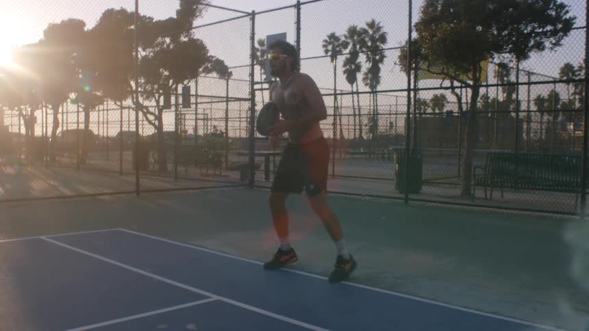 An athletic man plays a fastpaced game of pickleball on an outdoor court the setting sun casting a warm flare over the surrounding palm trees