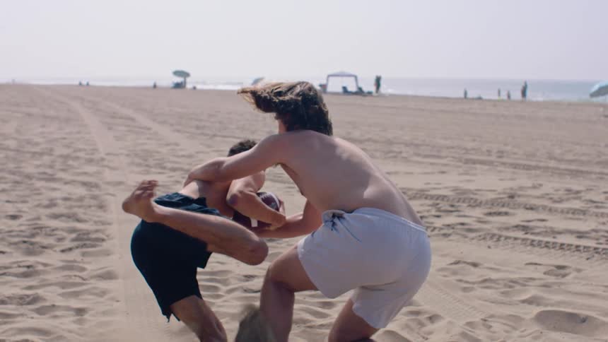 Two teenage boys playing rugby on the beach at midday with one tackling the other to the ground on a sunny day