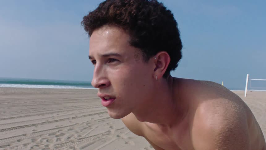 The face of a shirtless young man on a beach holding an american football ball
