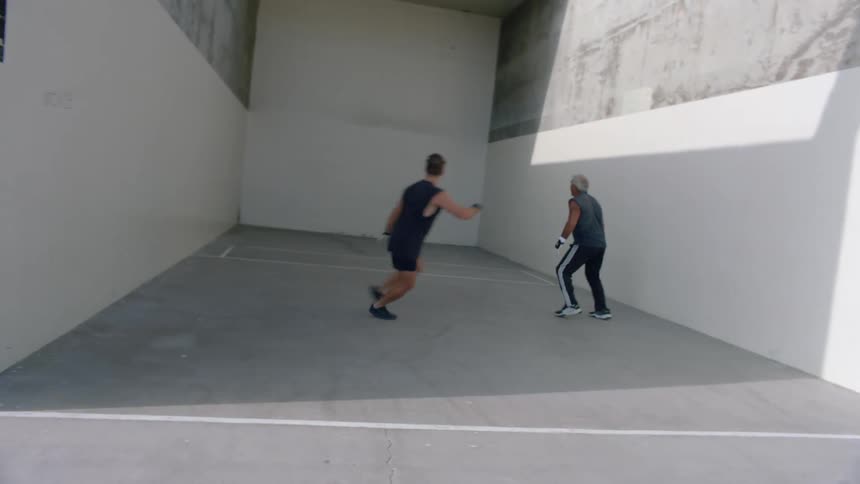 Two men engage in a dynamic handball match hitting the ball against the large white walls of an outdoor court on a bright sunny day