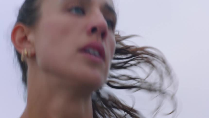 A woman with blue eyes and gold jewelry is seen in a closeup shot as she appears to be running or moving quickly her face shows focus and physical exertion against a blurred lightcolored background