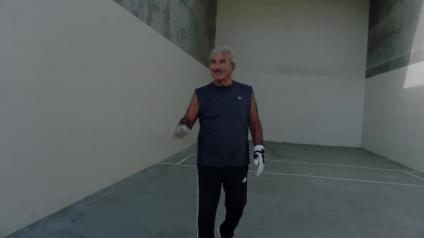 A man with white hair and a black tank top is wearing gloves in an environment surrounded by large walls
