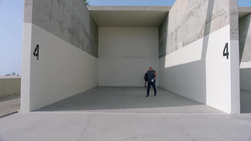 Two men are using gloves to hit balls against a wall
