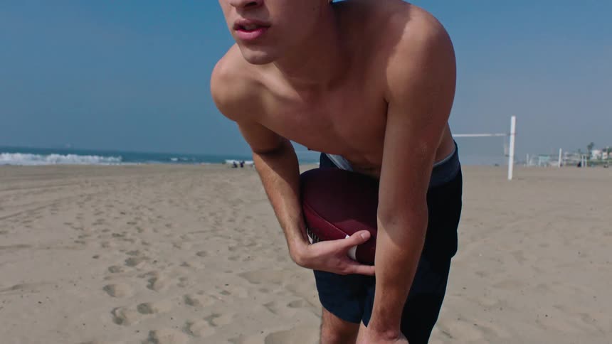 Young man leaning over on the beach on a sunny day holds a football