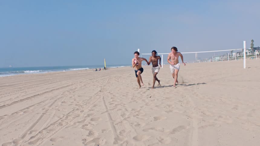 Three shirtless young men race toward the camera across a wide sandy beach on a sunny day with one carrying an american football while his two friends chase him past a volleyball net with the blue ocean