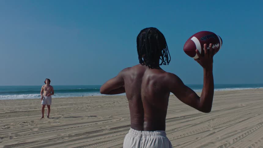 Two shirtless men wearing shorts on beach sand one throwing a football to the other on a sunny day