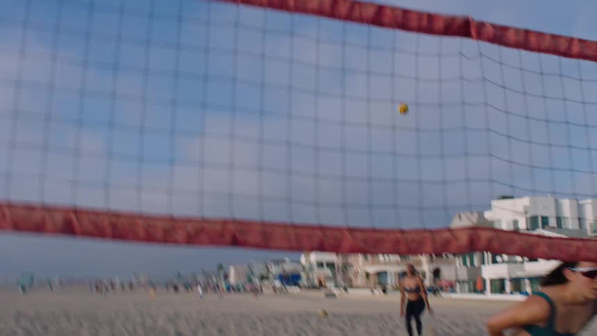 A woman is crouching behind a volleyball net while someone behind her is serving a ball
