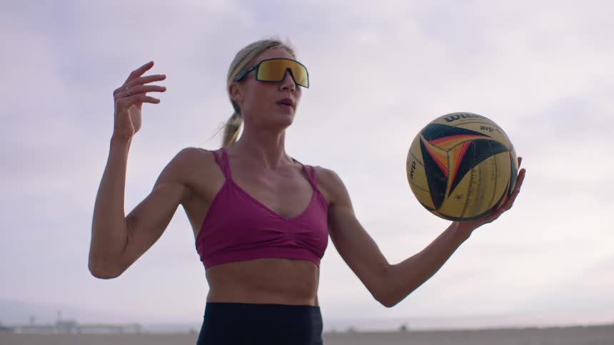 A woman wearing a pink top and yellow sunglasses is holding a volleyball in her hands and spinning it