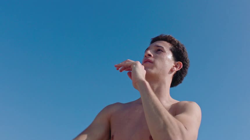 A young man outdoors shirtless throwing a football to another person under bright daylight