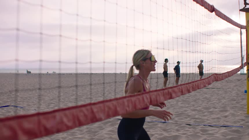 A blonde woman wearing glasses is behind a volleyball net walking on the beach sand