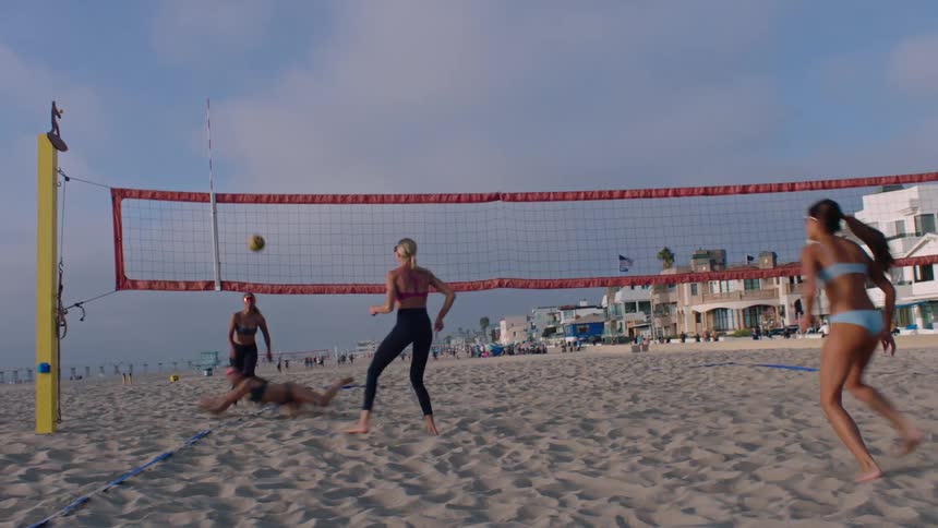 Women play volleyball on the beach near a row of apartment buildings on a summer day