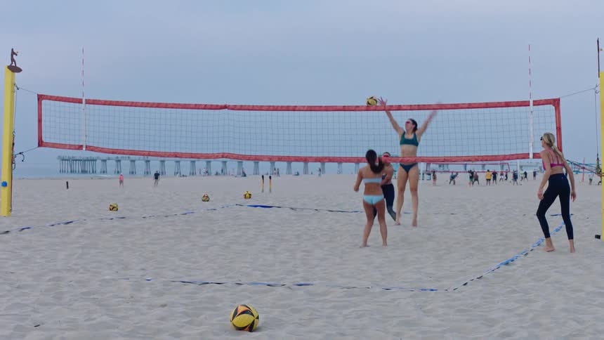 Four women play beach volleyball on a sandy court where one player serves the ball over the net and the opposing team attempts to return it during an overcast day with other people on the background