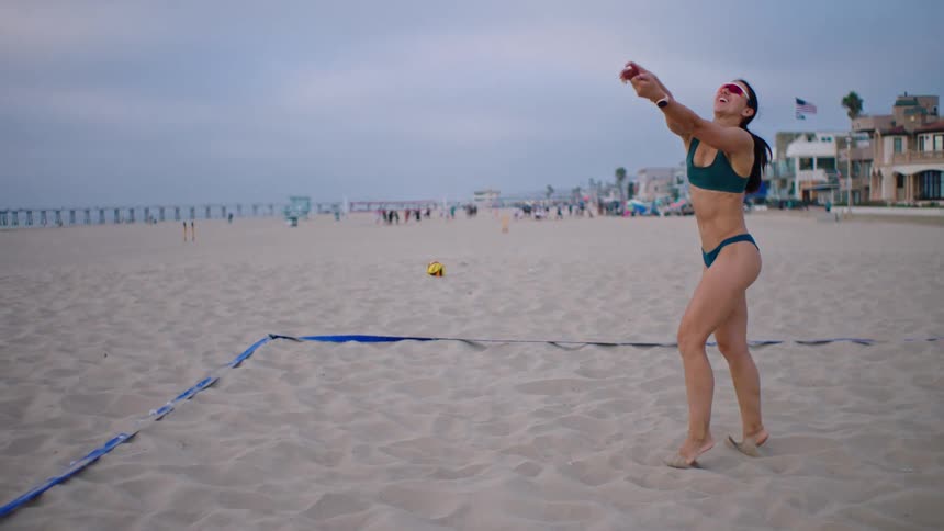 Young woman in a bikini wearing sunglasses playing volleyball on the beach at midday