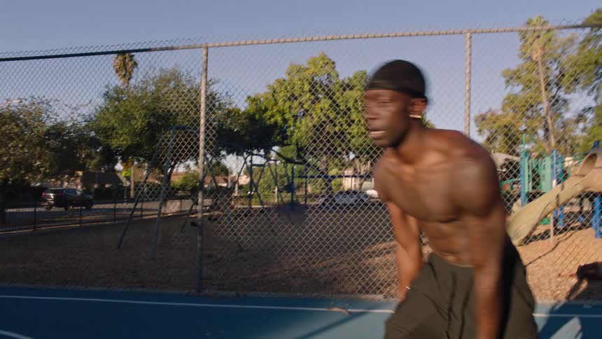 A shirtless muscular man wearing a black doo rag is intensely dribbling and shooting a basketball on an outdoor court next to a chain link fence and park