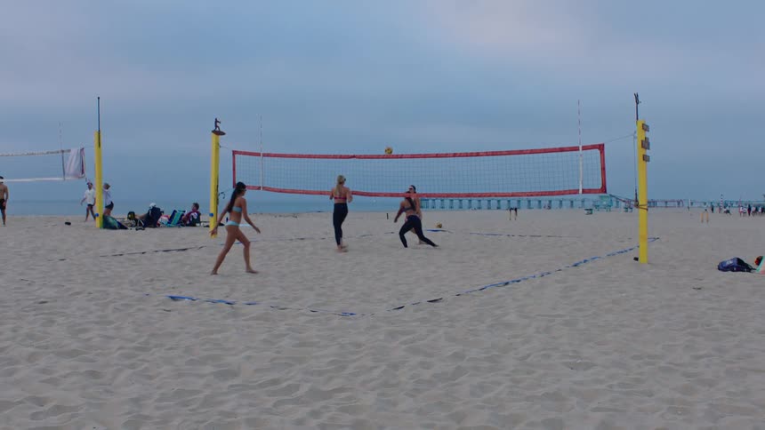 A group of people plays beach volleyball on a sandy court under a cloudy sky one player serves the ball over the net while others move into position to react as it falls into the sand