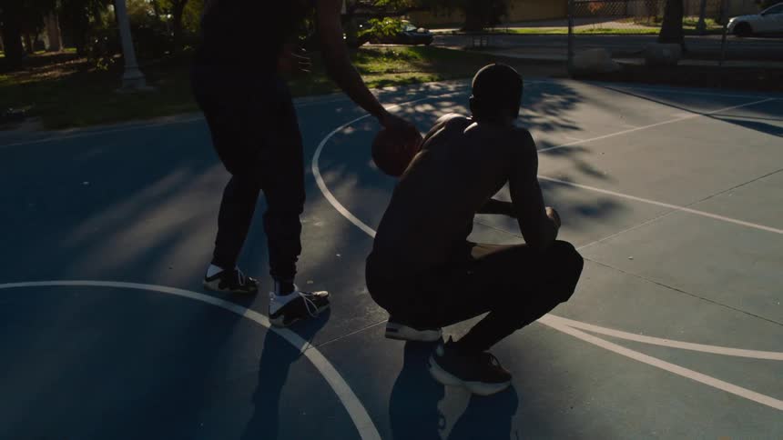 A man is crouching on a basketball court while a man next to him is dribbling a ball on the ground
