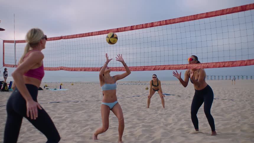 A group of young women playing volleyball on the beach all wearing sunglasses on a cloudy day