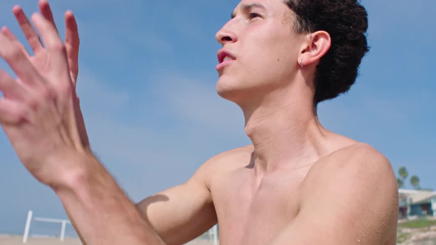 A shirtless young man stands on a sunny beach and catches a football thrown from offcamera before preparing to toss it back against a backdrop of palm trees and coastal homes