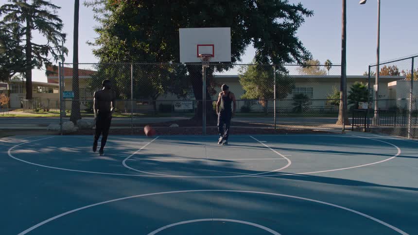 Two people are playing an intense oneonone basketball game on an outdoor blue court in a sunny neighborhood park with one player dribbling aggressively toward the hoop as the other closely defends near the basket framed by tall trees and a fence