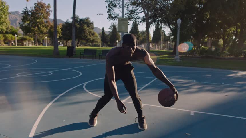 A shirtless man in black leggings and a skull cap performs rhythmic crossover dribbles with a basketball on a blue outdoor court while sunlight filters through the surrounding park trees and mountains in the distance