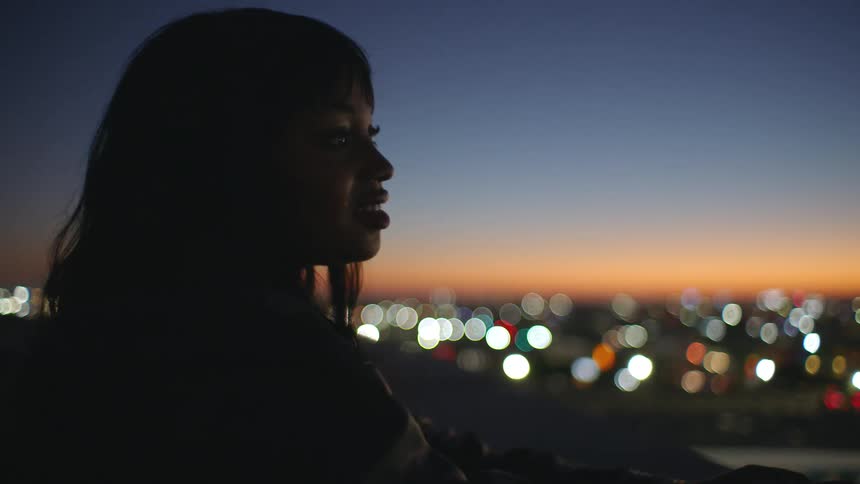 Young woman standing on a rooftop at night smiling as she looks out over the city with blurred lights illuminating the buildings behind her