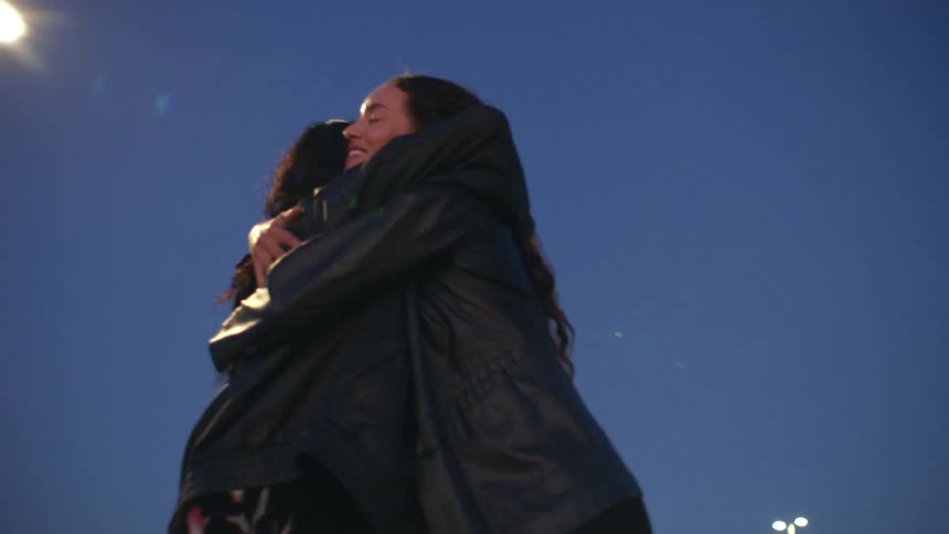 Two women jump with joy and share an enthusiastic embrace against a deep blue night sky on an outdoor rooftop illuminated by a nearby street lamp