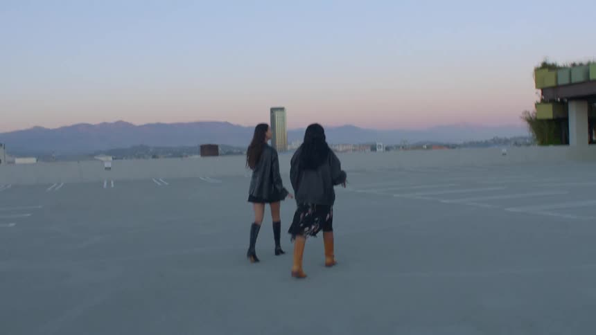 Two women wearing jackets and boots walk away from the camera across an empty rooftop parking lot toward a city skyline and mountains during dusk
