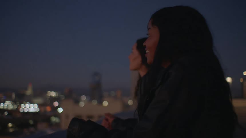Two female friends conversing while standing on a rooftop in a city at night
